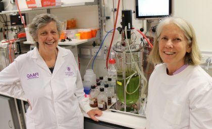 Professor Mary Fletcher and Diane Ouwerkerk with the fermenter which mimics conditions in a cow's stomach and will be used to test the biopolymer. two women in white coats smile in a laboratory in front of a device containing green liquid